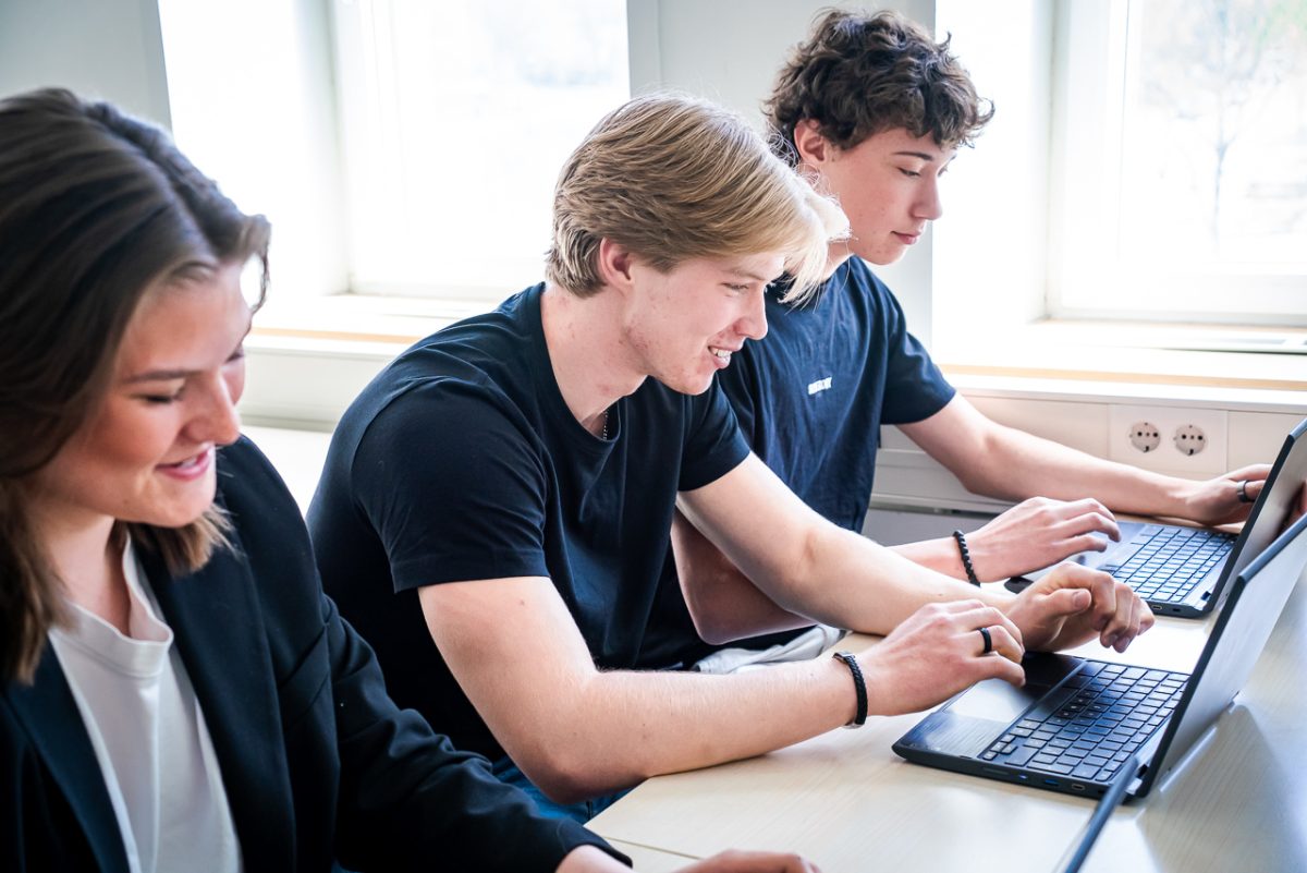 Tre elever sitter vid skolbänken och arbetar på sina datorer.
